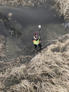 Harper Baldwin stands at the base of a hill with sand and dried grass. She is holding a Trimble instrument. 