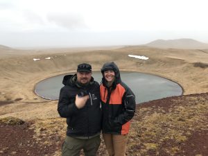 Alex Zaochney and Harper Baldwin stand next to each other for a photo. Behind them is a circular lake at the base of some rolling hills. There is dried grass on the hills. The sky is overcast and misty.