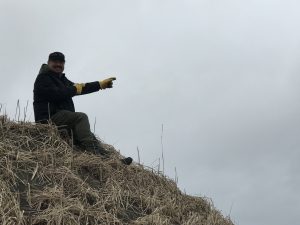 Alex Zaochney sits on top of a hill pointing in front of him. The hill is covered in dried grass and sand. The sky is overcast.