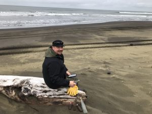 Alex Zaochney sits on a piece of driftwood on a sandy beach. 