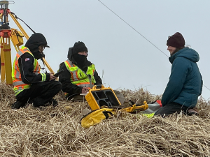Chris Tran, Chris Maio, and Harper Baldwin sit on dried grass near a set-up Trimble base. The sky is overcast and the three of them are wearing thick jackets.