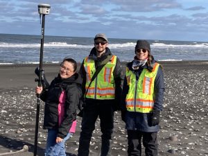 Someone stands next to Reyce Bogardus and Harper Baldwin, holding a Trimble instrument. They are standing on a semi-rocky beach. There's some white water on the crest of small waves. The sky is cloudy.