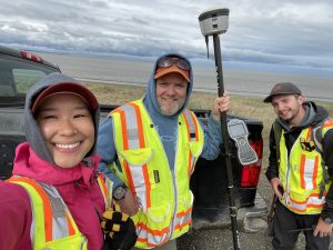 Roberta Glenn, Chris Maio, and Reyce Bogardus stand near each other around the bed of a black truck. Chris is holding the Trimble instrument. The ocean can be seen in the background and the sky is overcast.