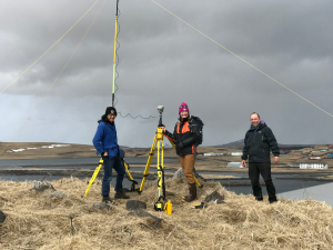 Chris Tran, Harper Baldwin, and Matthew Balazs stand around a setup Trimble base with a tall antenna. They are standing on a hill with dried grass and a few jagged rocks. Behind them is a water body with smooth water and a few buildings from the nearby village. The sky is overcast.