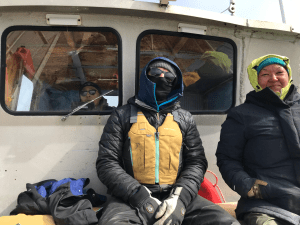 Someone sits inside the bridge of a boat, looking out the window. In front of the window sit two people, smiling at the camera. 