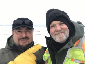 Alex Zaochney and Chris Maio stand next to each other for a selfie. The background is mostly whited out, but the horizon and a few rocks are barely visible. 