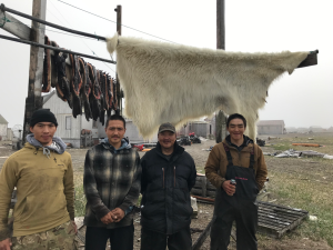 Four locals from Gambell stand next to each other. Behind them is a large polar bear skin and a row of seal skins drying on racks. Further back are a few buildings from the village, some ATVs and some debris around the patchy ground. The sky is overcast and foggy.