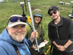 Chris Maio, Seth Classen, and Reyce Bogardus stand near each other for a selfie. Chris is holding the Trimble equipment and Seth is holding the field notebook. They are standing in a field of green grass. There is some debris in the background.