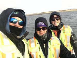 Chris Maio, Seth Classen, and Harper Baldwin stand together for a selfie. They seem to be standing on a vessel in a large body of water. A strip of land can be seen in the background. The sky is overcast.