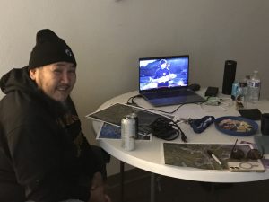 Someone sits at a table with various maps strewn about. There is a video playing on a nearby laptop. They appear to be indoors.