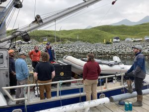 A group of people stand around a water vessel. Some are on the vessel and a couple are standing on the nearby dock. They are in a harbor. There are cars parked behind the rip-rap. There are green hills further back. The sky is overcast.