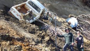 Reyce Bogardus and Roberta Glenn stand next to a tall bluff with debris strewn about. Among the debris is a rusted truck. Reyce is holding the Trimble staff and Roberta is holding the Trimble controller.