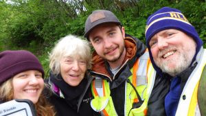 Jessica Christian, Sue Flensburg, Reyce Bogardus, and Chris Maio stand around each other for a group selfie. Behind them is vibrant green brush.